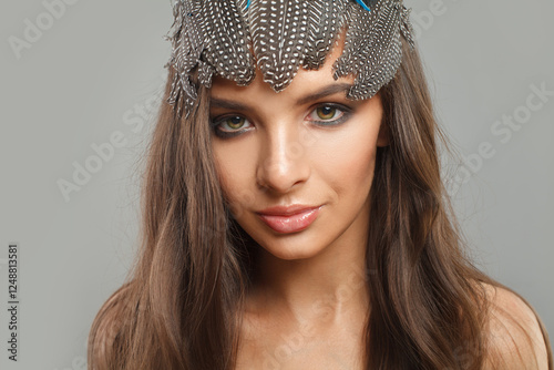 Charming female model with healthy long wavy hairstyle, trendy make-up and hairdeco feathers accessories on her head looking at camera, fashion beauty portrait photo