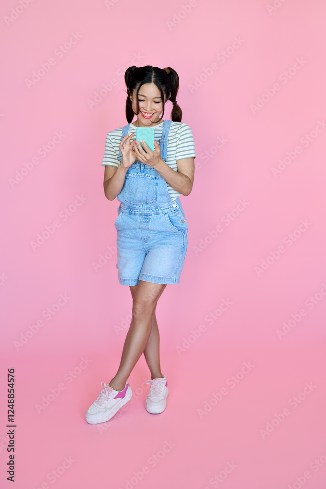 Smiling happy young Asian woman using mobile phone doing ecommerce shopping in smartphone apps, playing games, checking social media standing isolated on pink background. Full body length photo. Smiling happy young Asian woman using mobile phone doing ecommerce shopping in smartphone apps, playing games, checking social media standing isolated on pink background. Full body length photo.