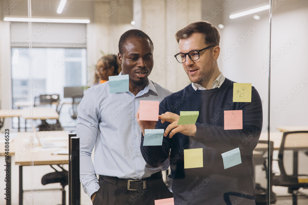 Two businessmen discussing ideas with sticky notes on a glass wall in a modern office, showcasing teamwork and collaboration Two businessmen discussing ideas with sticky notes on a glass wall in a modern office, showcasing teamwork and collaboration