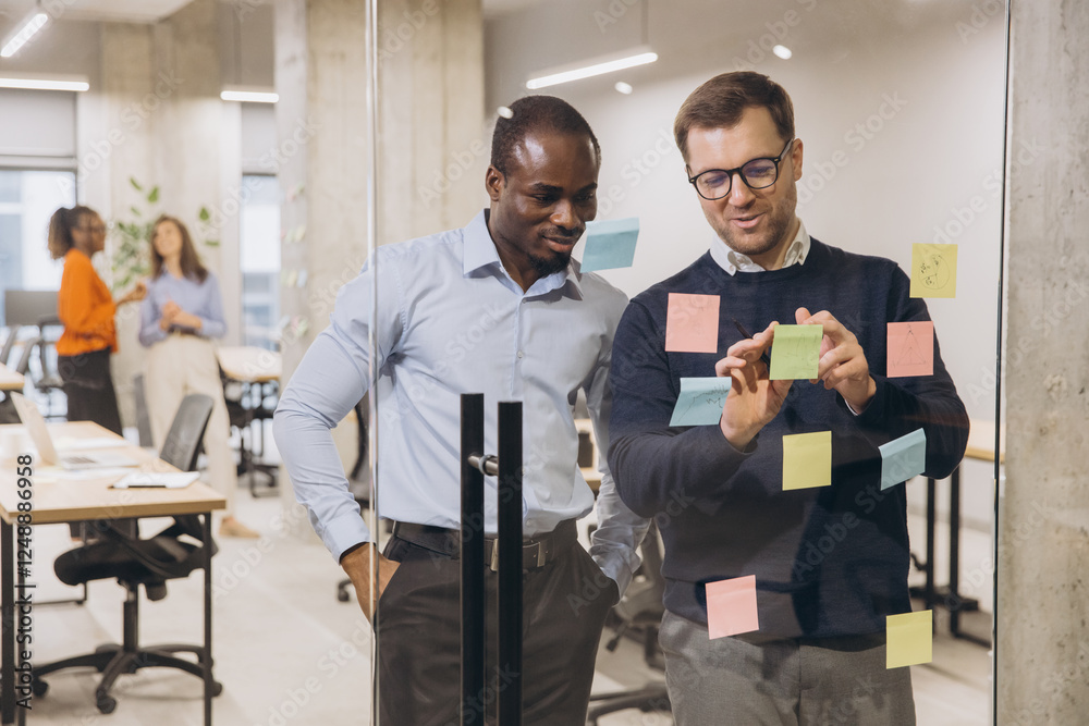 Two diverse businessmen collaborating on a project, using sticky notes on a glass wall in a modern, open plan office setting Two diverse businessmen collaborating on a project, using sticky notes on a glass wall in a modern, open plan office setting