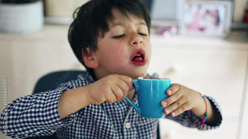 Young boy drinking hot chocolate for breakfast from a blue mug, with an expressive face and playful demeanor in a bright indoor setting Young boy drinking hot chocolate for breakfast from a blue mug, with an expressive face and playful demeanor in a bright indoor setting