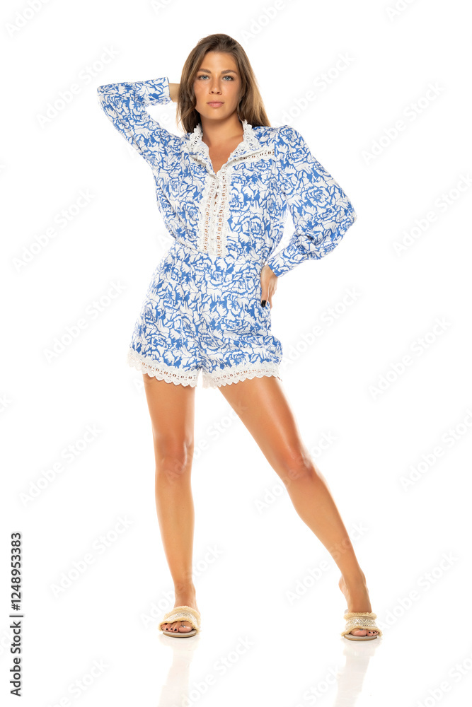 Young woman in blue and white floral romper with lace trim. Long brown hair, tanned skin. Full-body shot against white background. Wearing slide sandals Young woman in blue and white floral romper with lace trim. Long brown hair, tanned skin. Full-body shot against white background. Wearing slide sandals