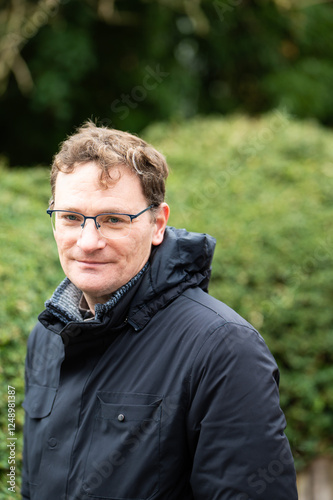 Portrait of a 45 yo white male wearing a dark winter jacket against a  green nature background, Koekelberg, Brussels, Belgium