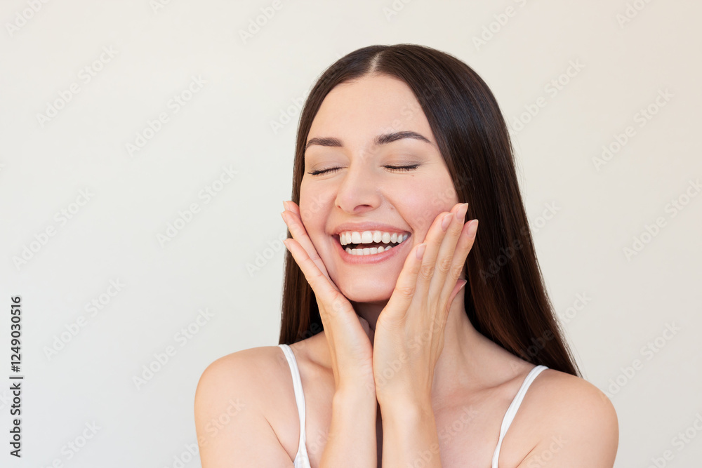 Fototapeta premium A young pretty Caucasian brunette woman smiling, closing her eyes and gently touching her face with her hands. 