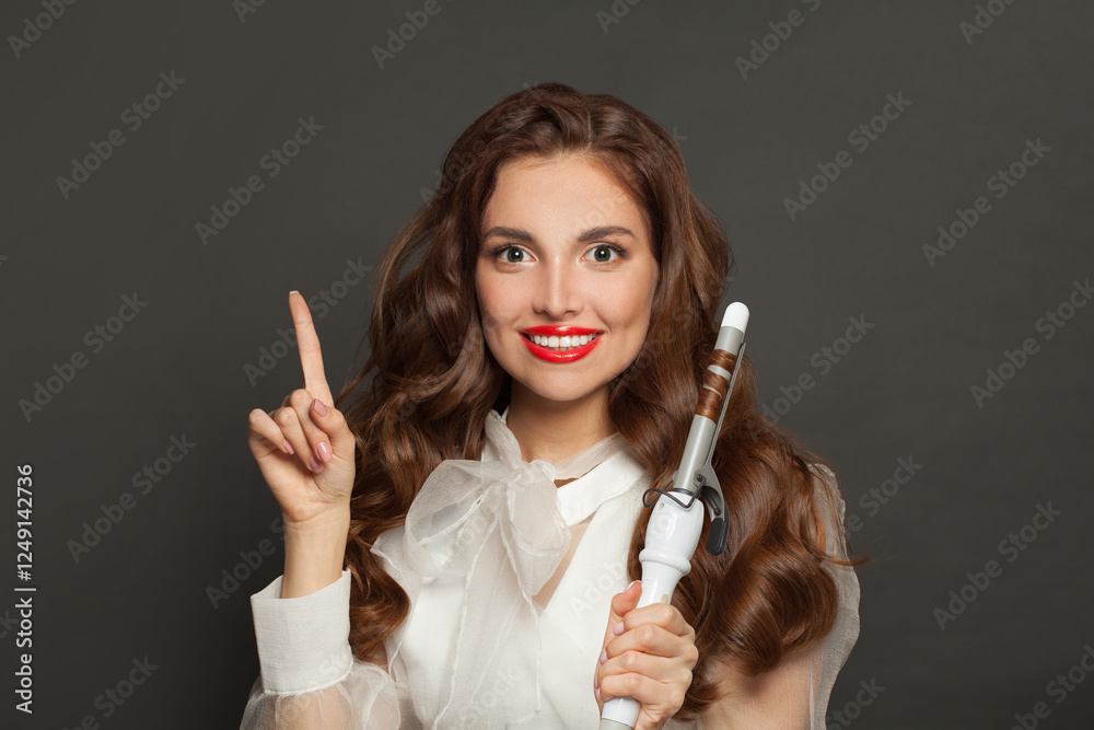Obraz premium Attractive woman with healthy long wavy hair pointing and using curlyng iron, fashion beauty portrait
