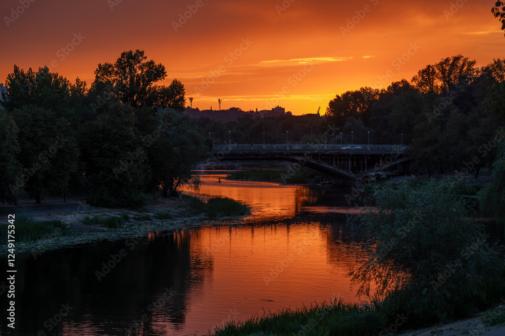 Fototapeta premium Stunning sunset over a calm river with a silhouette of a bridge and trees at twilight