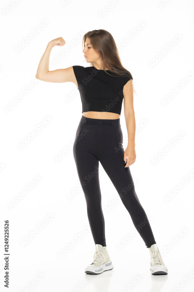 Fototapeta premium Woman in black activewear flexing bicep, showing strength and confidence on white background with white sneakers