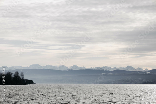 Wide view of a calm lake with mountain ranges in the distance under a cloudy, overcast sky. photo