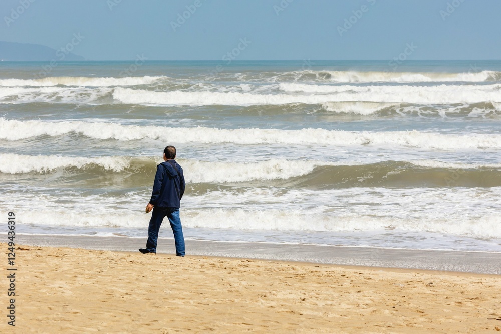 Adult male walking on sandy beach by ocean waves on a cloudy day Adult male walking on sandy beach by ocean waves on a cloudy day