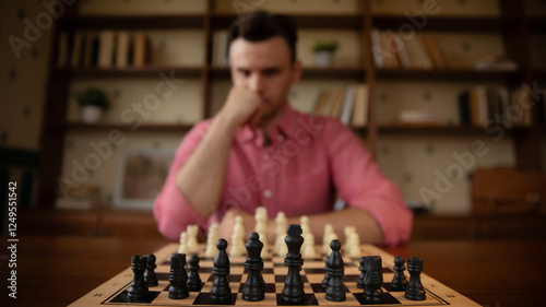 A man in a pink shirt thoughtfully ponders his next move in a chess game. The focus is on the black pieces in the foreground, with the blurred background creating a deep, reflective mood. photo