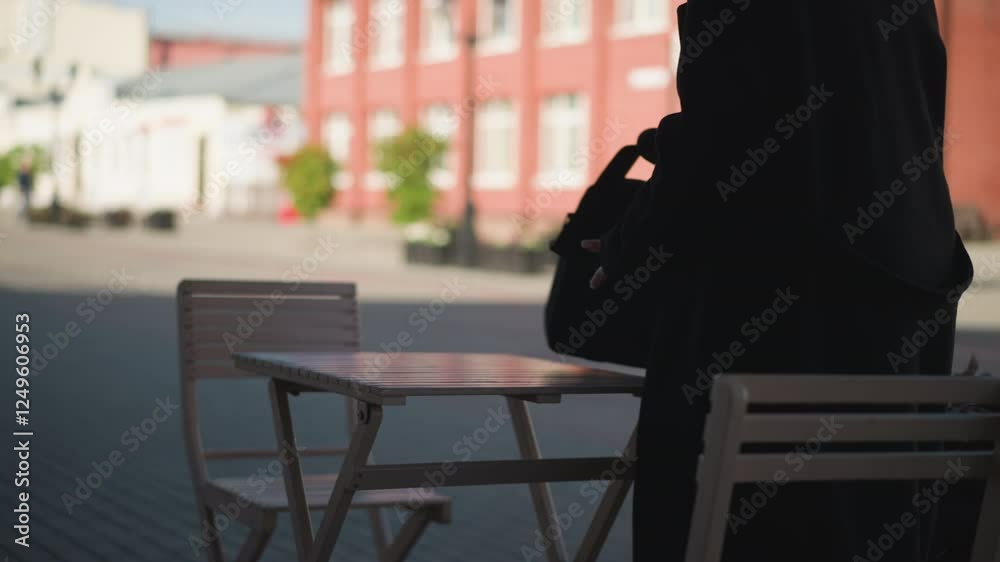 Back view of woman placing her bag on wooden table and sitting down at outdoor cafe with urban building and someone walking in blurred background Back view of woman placing her bag on wooden table and sitting down at outdoor cafe with urban building and someone walking in blurred background