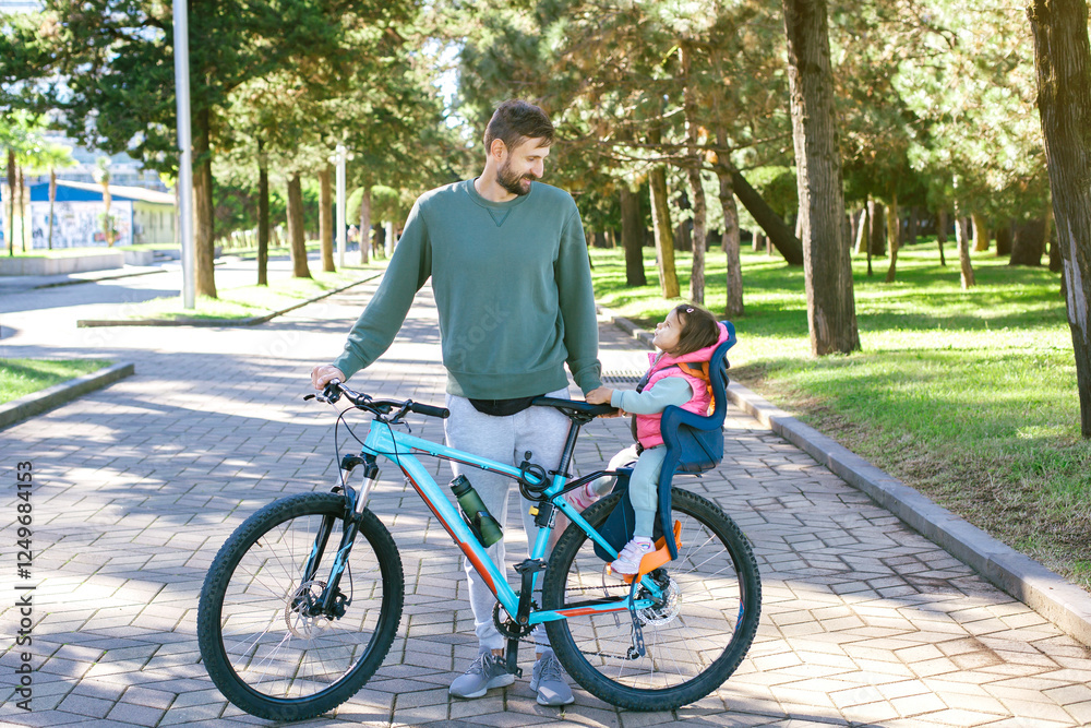 Father with a bicycle and his little daughter sitting in a child bike seat in a green park, smiling at each other, enjoying outdoor family time in sunny day. fatherhood concept Father with a bicycle and his little daughter sitting in a child bike seat in a green park, smiling at each other, enjoying outdoor family time in sunny day. fatherhood concept