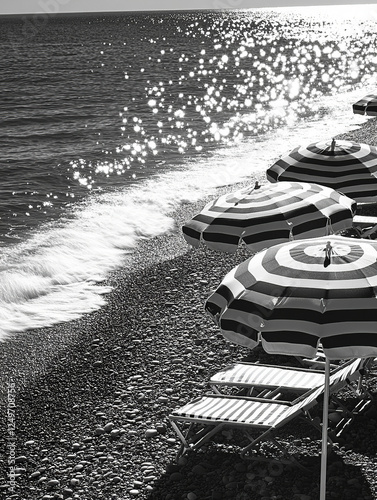 Create a detailed, enlarged, black and white photograph of a French Riviera beach scene. Focus on gentle waves lapping against soft pebbles and sand, with sunlight reflecting off the water in delicate