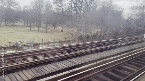 A train moves along the tracks, passing by a serene park with bare trees and a grassy area. The overcast sky adds a calm atmosphere to this peaceful setting