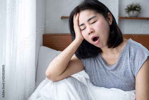 Asian woman waking up tired due to sleep inertia, yawning, and feeling fatigued in bed