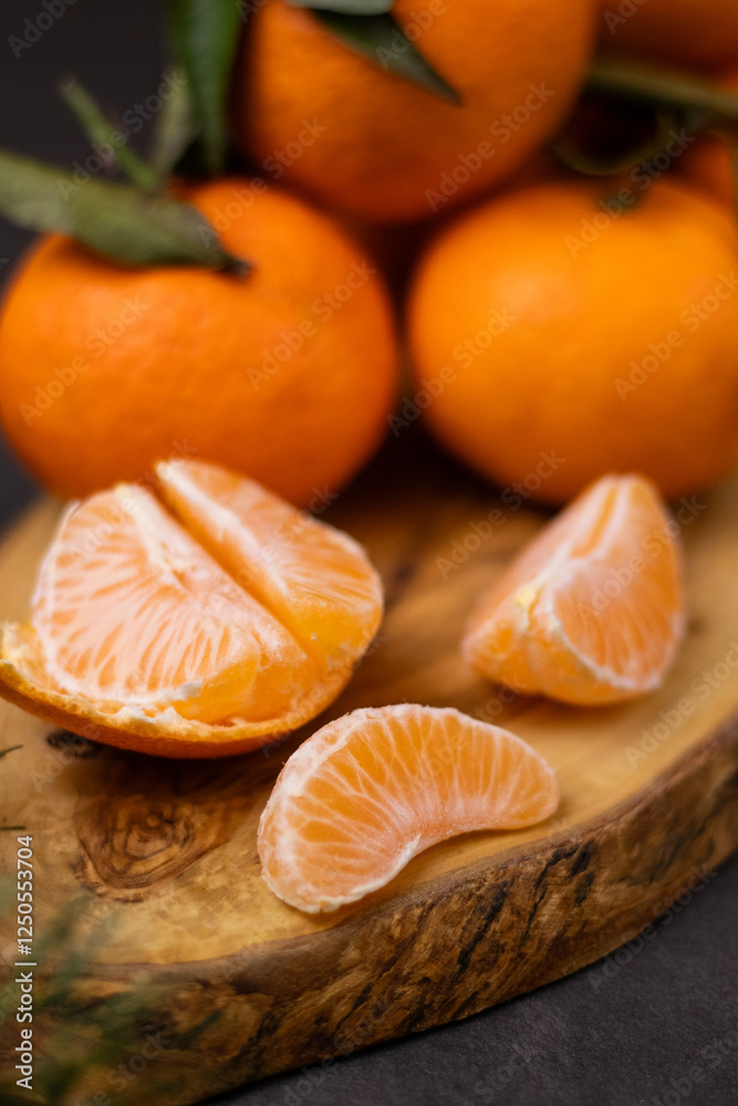 Close up of peeled mandarine slices and a pile whole mandarines on wooden plate, blurry green pine branch in front Close up of peeled mandarine slices and a pile whole mandarines on wooden plate, blurry green pine branch in front