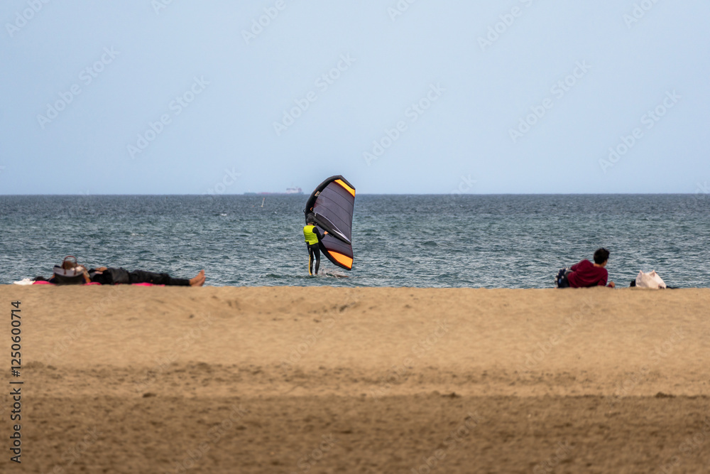 Wing foiling getting ready to surf on the beach with some waves on a cloudy day and people relaxing on the sand of Leucate beach, France. Wing foiling getting ready to surf on the beach with some waves on a cloudy day and people relaxing on the sand of Leucate beach, France.