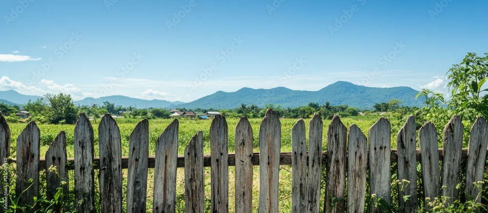 Fototapeta premium Rustic wooden fence framing a picturesque landscape in rural Thailand with mountains and clear blue skies in the background.