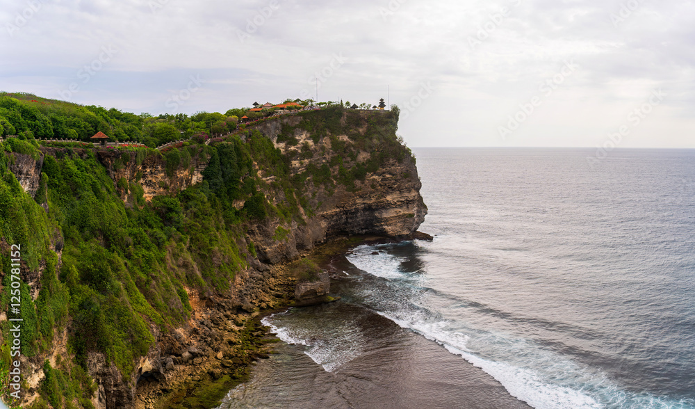 Nature's raw power meets spiritual serenity as waves crash against the base of a massive cliff crowned with Balinese temple architecture and lush vegetation