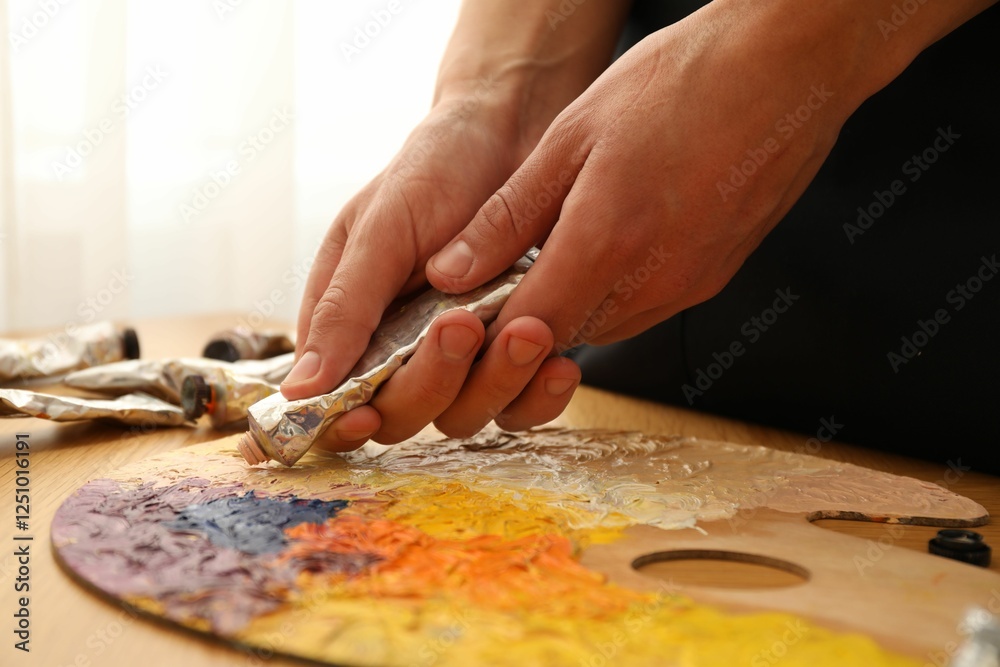 Man squeezing paint from tube onto palette at wooden table indoors, closeup Man squeezing paint from tube onto palette at wooden table indoors, closeup
