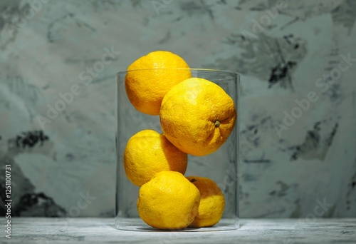 A cluster of vibrant yuzu lemons fills a transparent vase, set against a textured backdrop that enhances their sunny hue during daylight. The composition showcases their unique beauty photo