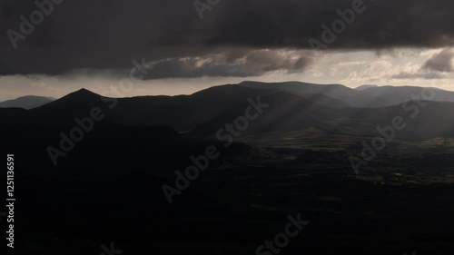 A dramatic landscape shows sun rays cutting through a cloudy sky over a series of hills and valleys. Lush green vegetation covers the lower slopes.