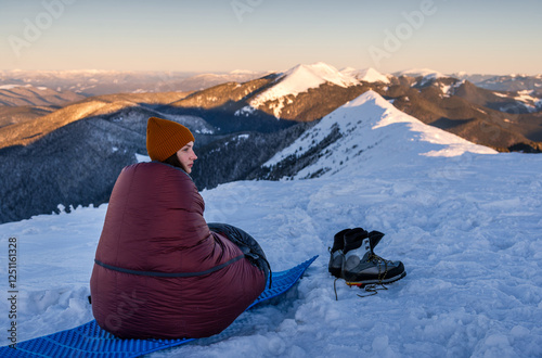 Young Woman in sleeping bag at winter mountains watching alpenglow sunrise. Bivouacs At Snowy Mountain Range. photo