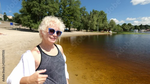 Blonde woman sixty years old in a white shirt walks near the river on the beach. Portrait of a woman in dark glasses. Beautiful old woman
