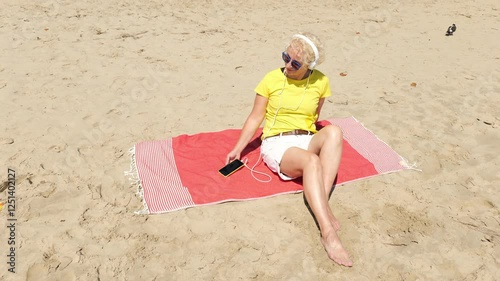 Beautiful cheerful sixty year old blonde woman listening to music in big white headphones sitting on the beach. Woman in yellow T-shirt and white shorts. Pensioner woman in sunglasses