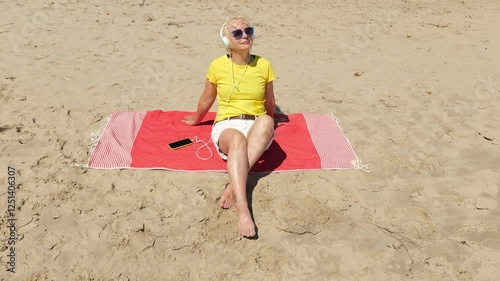 Beautiful cheerful sixty year old blonde woman listening to music in big white headphones sitting on the beach. Woman in yellow T-shirt and white shorts. Pensioner woman in sunglasses