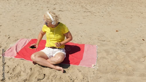 Beautiful cheerful sixty year old blonde woman listening to music in big white headphones sitting on the beach. Woman in yellow T-shirt and white shorts. Pensioner woman in sunglasses