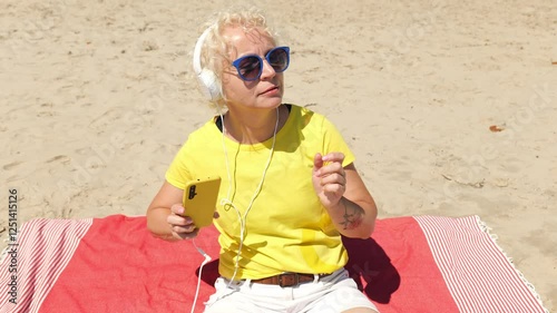 Portrait of a sixty year old blonde woman in sunglasses and a yellow T-shirt listening to music in big headphones while sitting on the beach. Woman cheerful old. Woman sportswoman