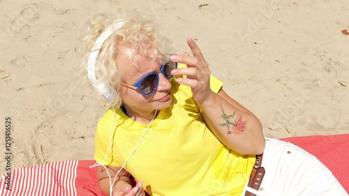 Portrait of a sixty year old blonde woman in sunglasses and a yellow T-shirt listening to music in big headphones while sitting on the beach. Woman cheerful old. Woman sportswoman
