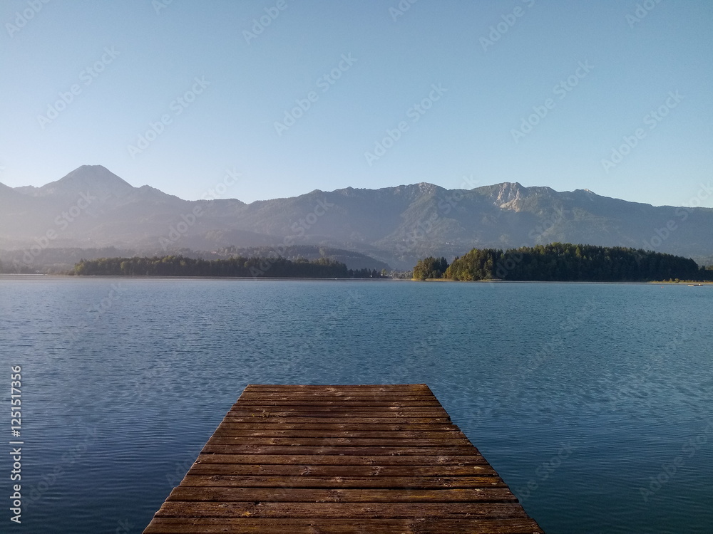 Fototapeta premium Panoramic view over beautiful lake Faak in Carinthia, Austria to Silhouette of Mittagskogel and Karawanks mountain range from wooden pier. Early morning in late summer.