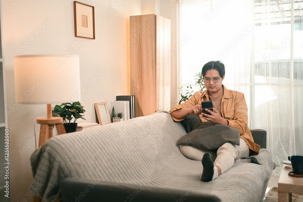 Fototapeta premium A man in casual wear relaxes on a cozy sofa, using his smartphone, Comfort and digital connectivity at home
