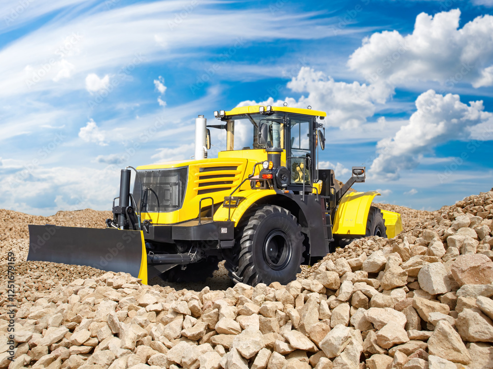 Obraz premium Bulldozer at stone quarry. Special equipment for mineral extraction. Yellow bulldozer on mountain of stones. Construction equipment. Wheeled bulldozer under blue sky. Development of stone deposit