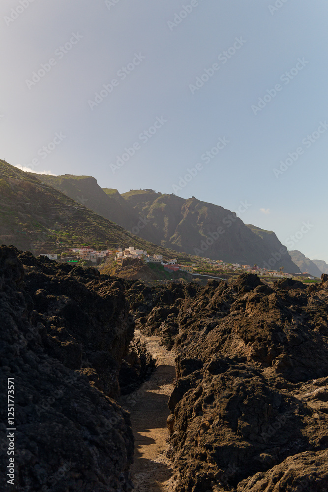 Obraz premium Man-made concrete footpath through volcanic rocky beach in Garachico, Tenerife. Vertical view towards the afternoon sun and coastal mountains.