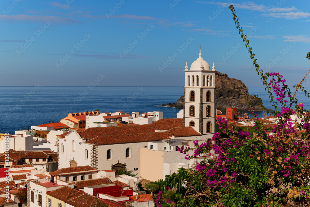 Above Garachico town in Tenerife with church and pink bougainvillea in front. Tall bell tower. Scenic Garachico rock island at the back. Calm sunny day with blue sky.