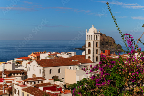 Obraz na płótnie Above Garachico town in Tenerife with church and pink bougainvillea in front. Tall bell tower. Scenic Garachico rock island at the back. Calm sunny day with blue sky. w salonie