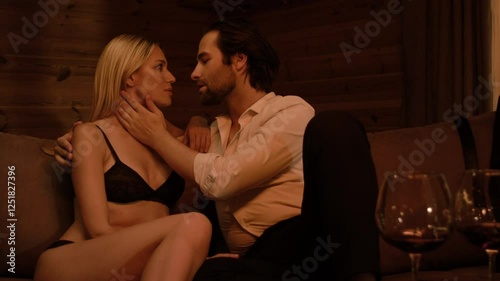 A stubbled brunette man in a white shirt and black pants gently caresses a blonde in black lingerie, their faces pressed close, lips almost touching. Two wine glasses complete the scene. photo