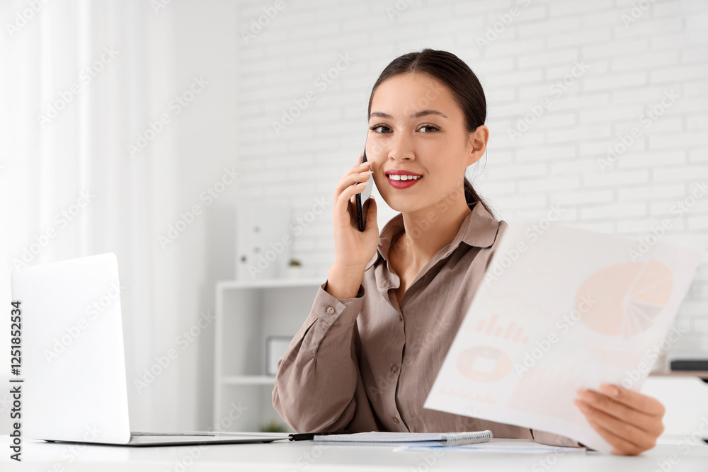 Young Asian businesswoman with charts talking by phone at her workspace in office Young Asian businesswoman with charts talking by phone at her workspace in office