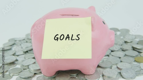 Goals written on a yellow note paper with a hand inserting coins into a pink piggy bank, surrounded by a pile of coins on a white background