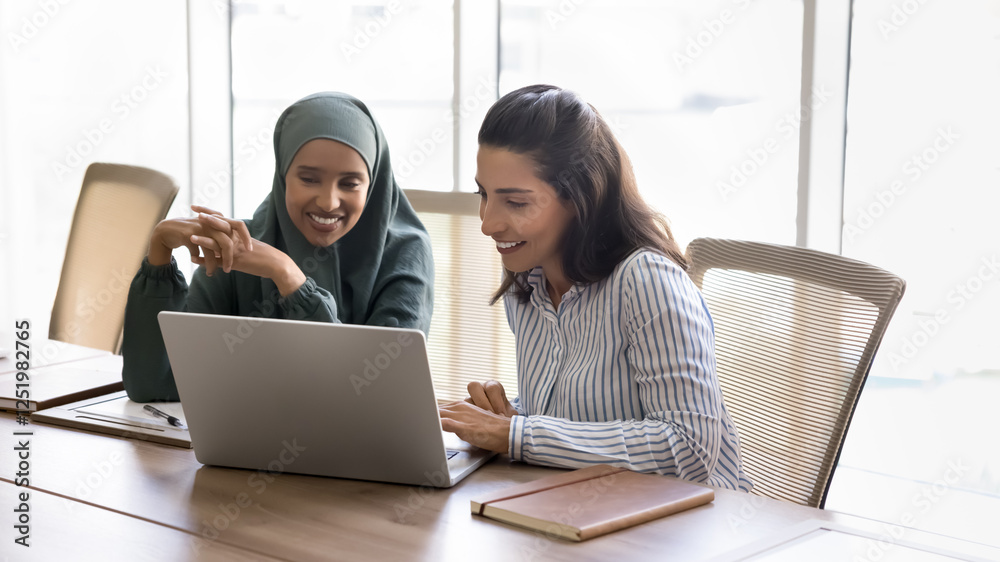 Young Muslim and Latin businesswomen, colleagues using laptop in office, going over presentation or preparing slides for meeting, view updates of project, check financial reports or budget allocations Young Muslim and Latin businesswomen, colleagues using laptop in office, going over presentation or preparing slides for meeting, view updates of project, check financial reports or budget allocations