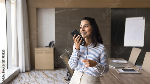 Smiling Brazilian businesswoman stands in modern conference room, holding smartphone recording voice message or lead conversation on speakerphone. Business, remote communication, voice recognition, AI