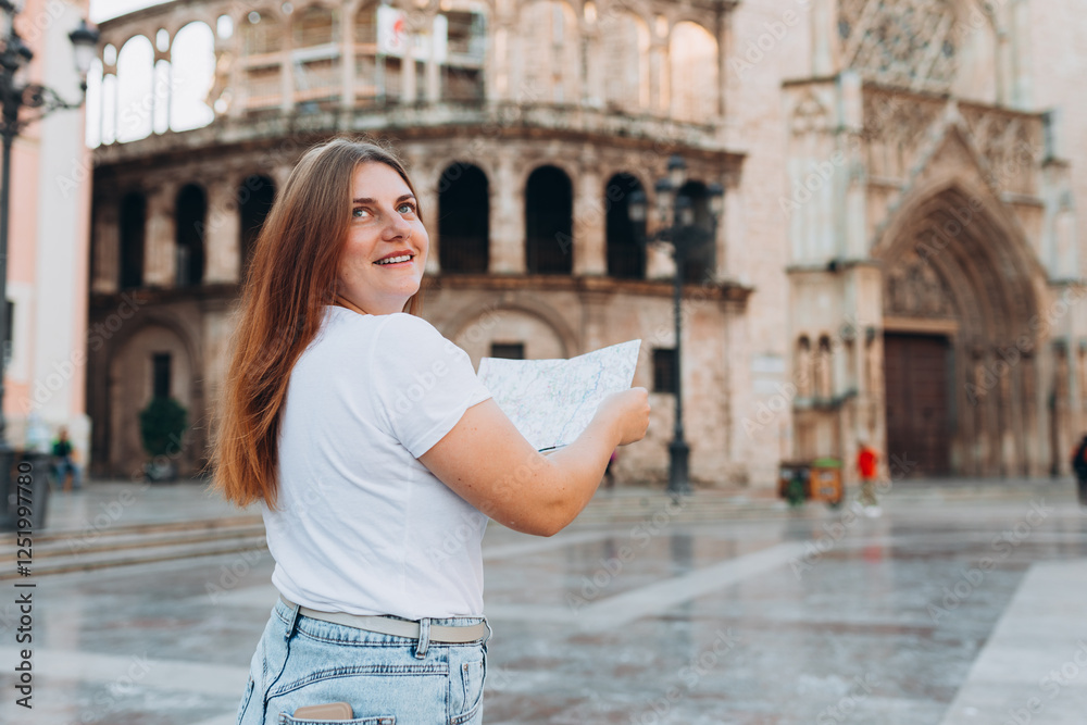 Portrait of a happy young woman with paper map walking in the city. Traveler 30s girl visiting Valencia Cathedral. Summer Holidays in Spain. Attractive female is exploring city. Top tourist attraction