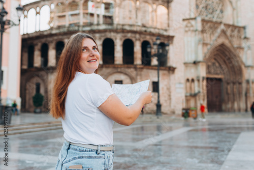Portrait of a happy young woman with paper map walking in the city. Traveler 30s girl visiting Valencia Cathedral. Summer Holidays in Spain. Attractive female is exploring city. Top tourist attraction