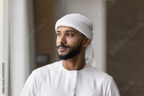 Confident Middle Eastern man stands indoor, gazing thoughtfully into distance. Young Arabian businessman, entrepreneur dressed in traditional white dishdasha, looking aside with pensive expression photo