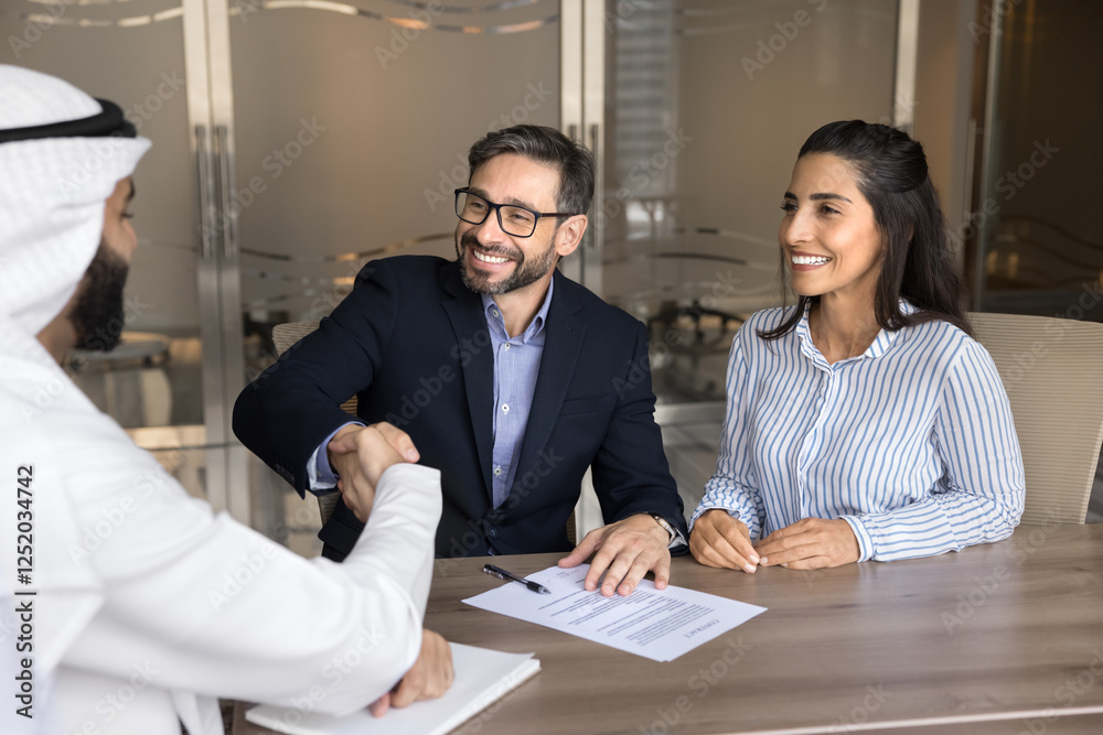 Manager and clients sealing professional agreement with handshake. Happy European spouses signing contract, shaking hands with Arabian male professional, express gratitude for getting services. Deal Manager and clients sealing professional agreement with handshake. Happy European spouses signing contract, shaking hands with Arabian male professional, express gratitude for getting services. Deal
