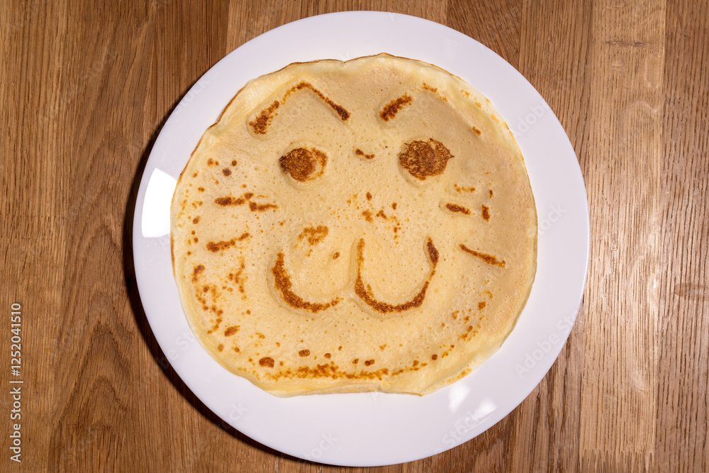 The image shows a delightful pancake cooked on the white plate, featuring a playful face with eyes, a nose, and a wide, smiling mouth. Kids meal cooking.