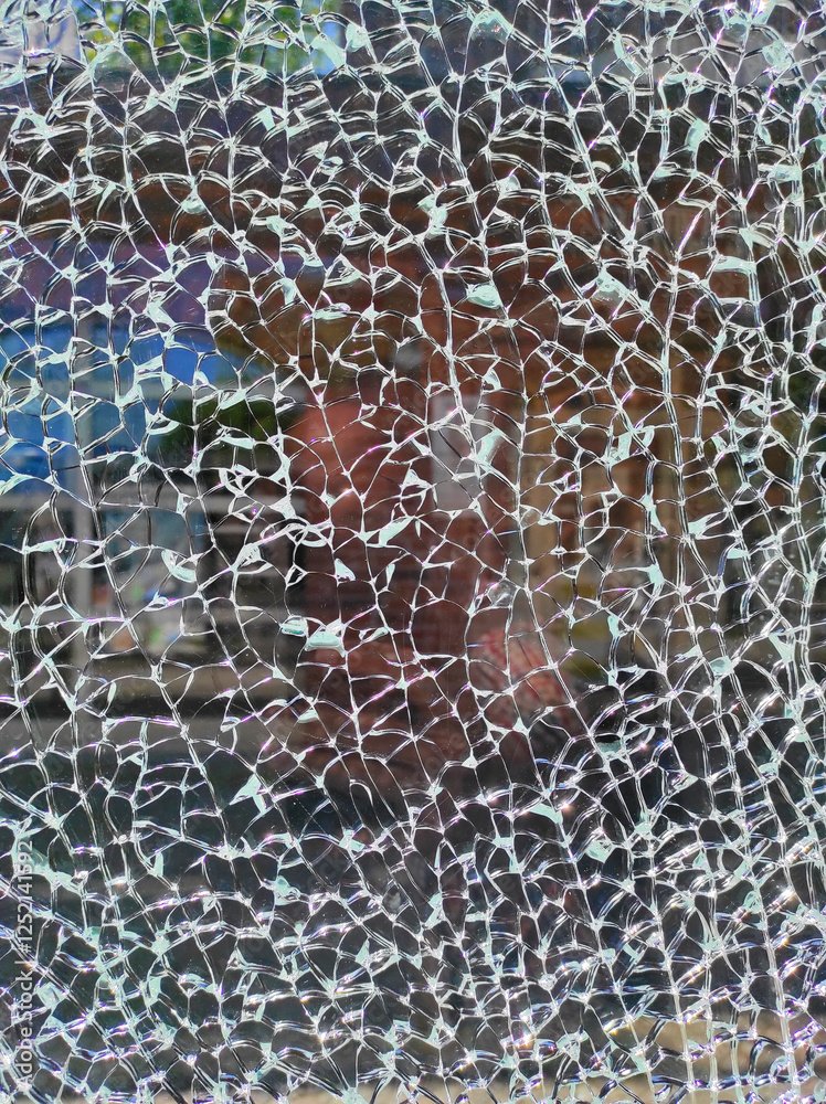 A close-up view of shattered glass reveals a distorted reflection of a nearby storefront, with sunlight casting intricate patterns through the cracks